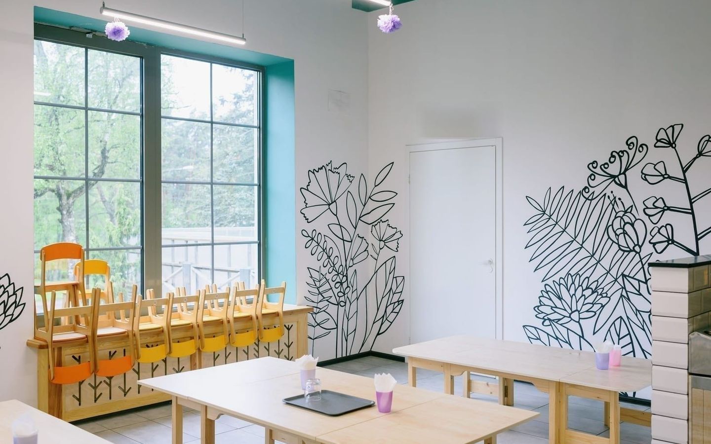 Bright room with child-sized chairs stacked by tall windows and botanical line-art painted on white walls