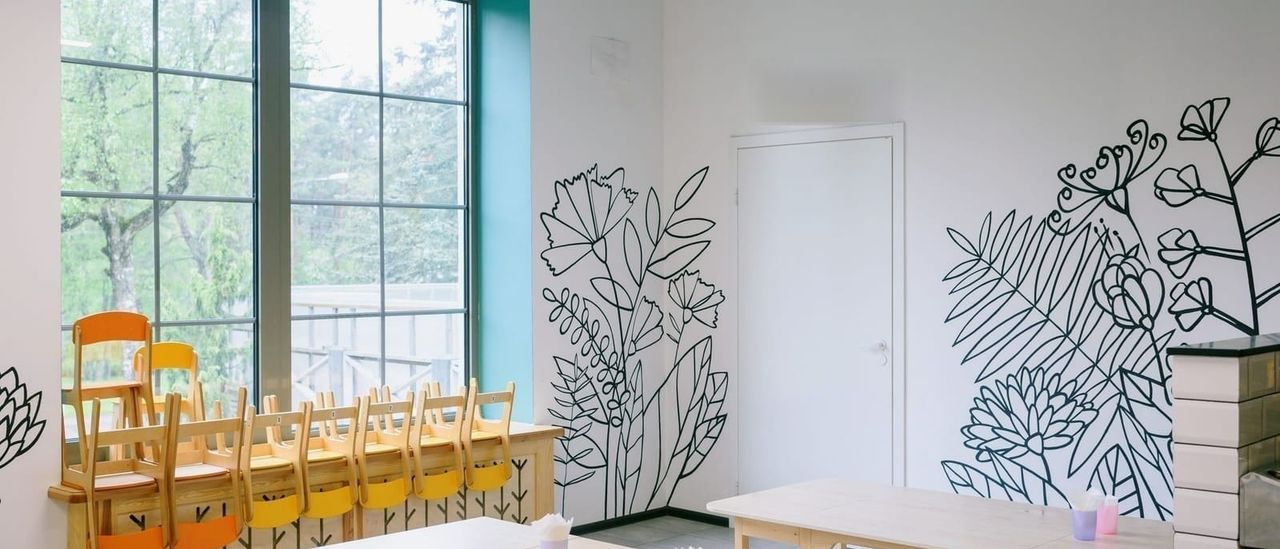 Bright room with child-sized chairs stacked by tall windows and botanical line-art painted on white walls
